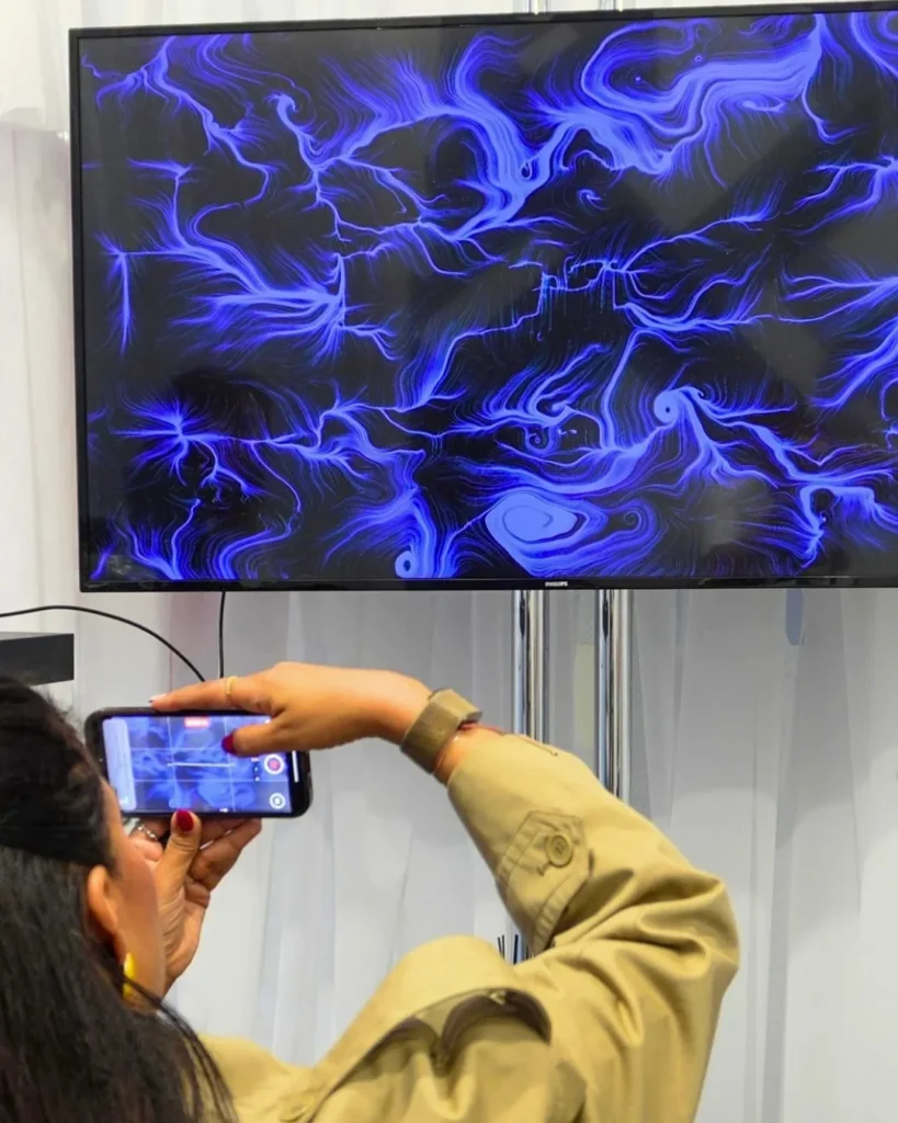 Una donna di spalle fotografa con lo smartphone uno schermo appeso a parete che mostra un'opera dalle linee e dai vortici blu e azzurri su fondo nero.