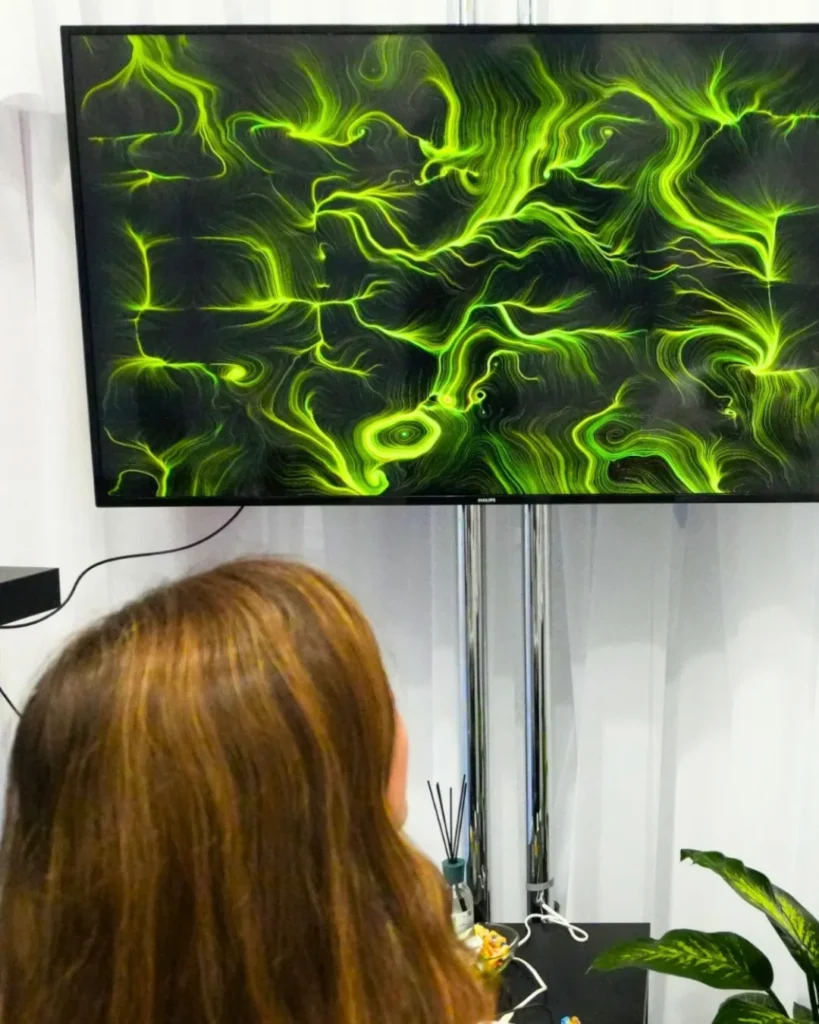 Una donna di spalle osserva uno schermo appeso a parete che mostra un'opera dalle linee e dai vortici verdi su fondo nero; sotto, su un piano scuro, alcuni bastoncini diffusori e una pianta.