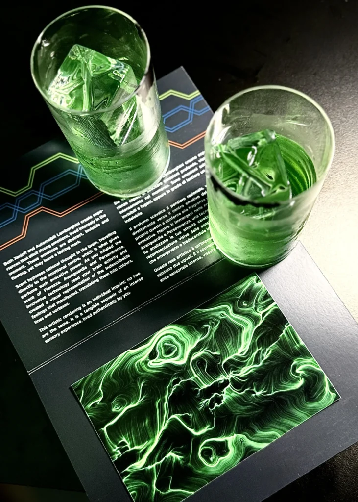 Due bicchieri con un cocktail verde e ghiaccio appoggiati su una cartolina aperta stampata; in basso una composizione astratta verde fatta di linee fluide.