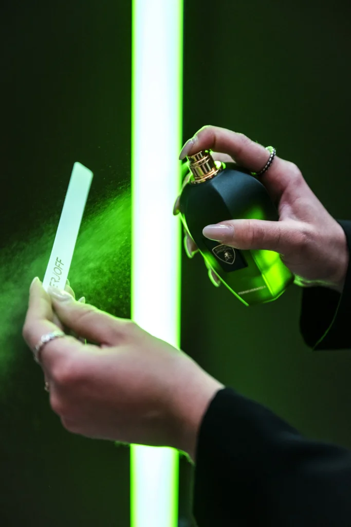 Due mani spruzzano una fragranza dal flacone Perseveranza di Xerjoff x Lamborghini su una mouillette, con la nuvola di profumo illuminata da una luce al neon verde.