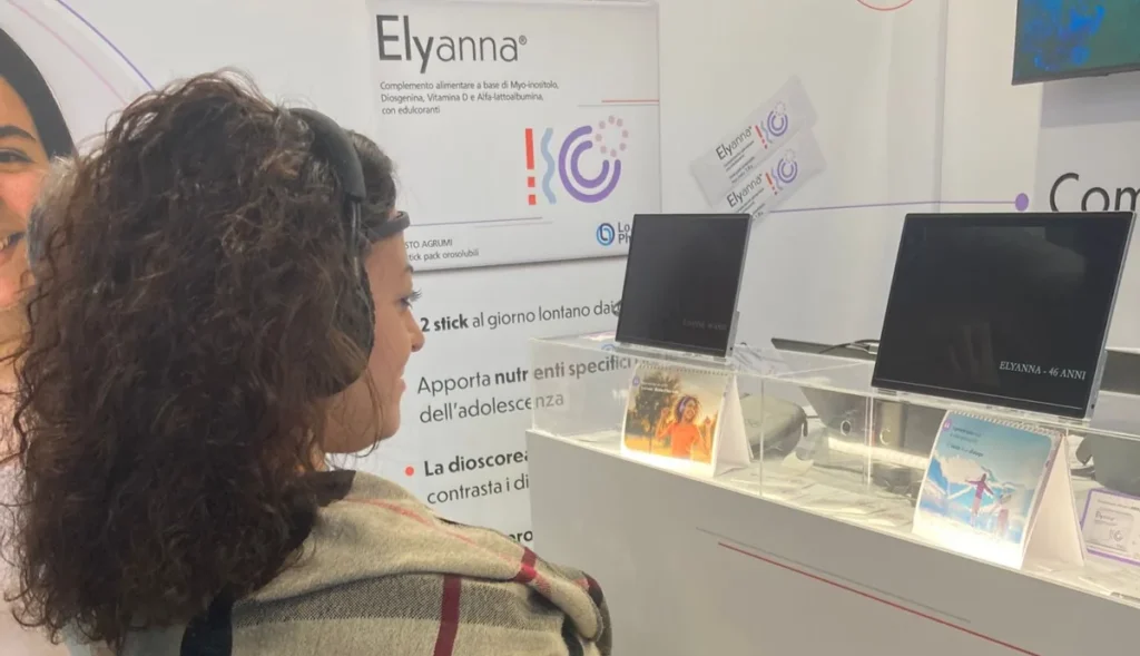 A participant with headphones and an EEG sensor on the forehead watches the Elyanna video in front of two screens at the Lo.Li. Pharma booth 