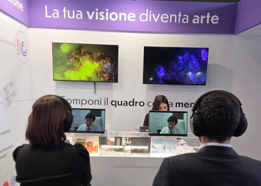 Two participants wearing headphones watch the Elyanna video at the Lo.Li. Pharma booth, above a green artwork and a purple one 
