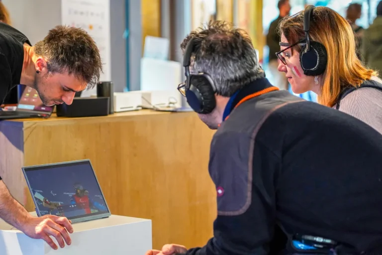 An operator checks the monitor while two participants wearing headphones watch the Milano Cortina Paralympics video